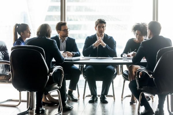 Confident businessman speaks diverse employees about project sit at around table in boardroom at company meeting. Leader discuss business strategy with colleagues. Mentor share thoughts at briefing.